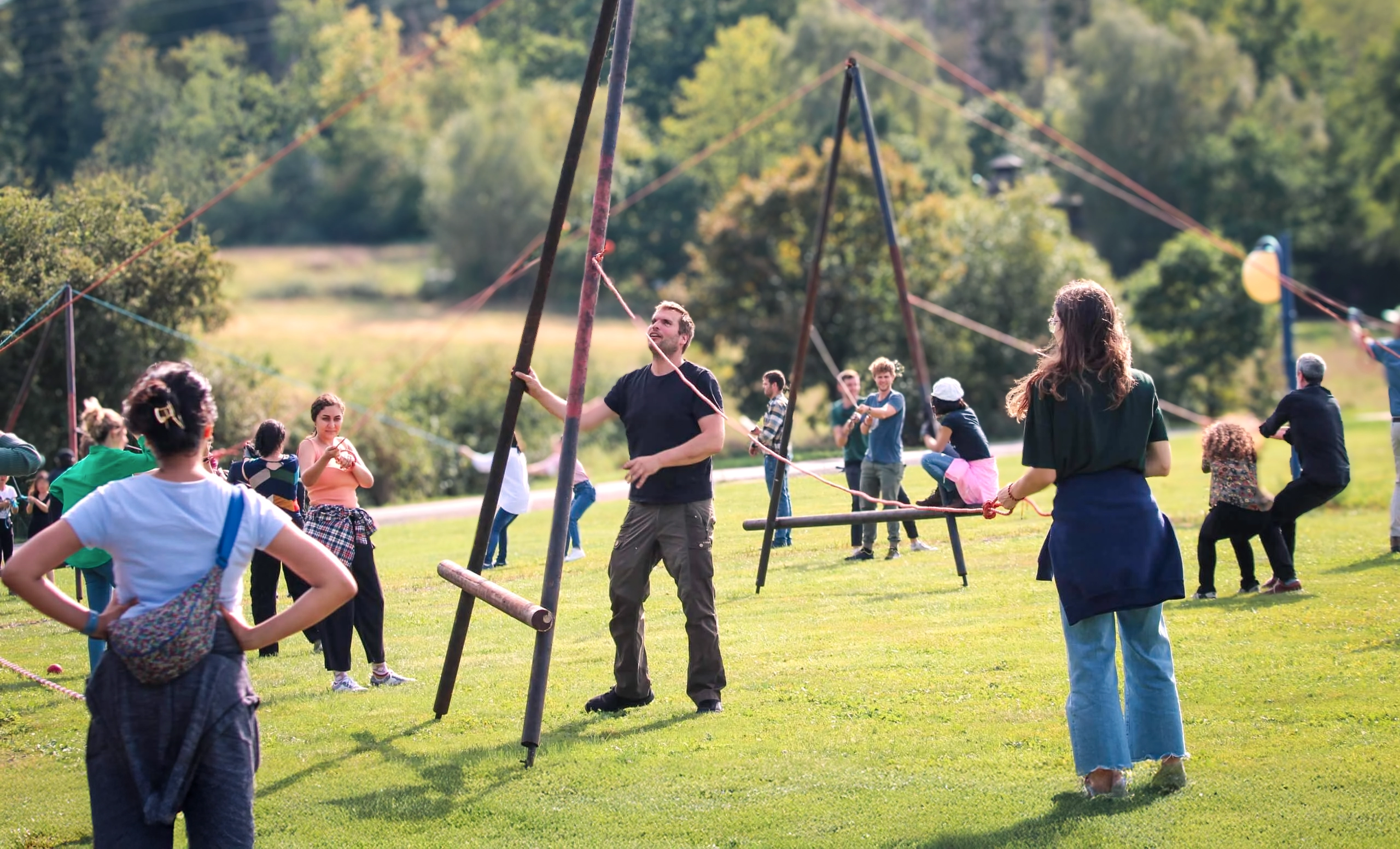 Eine Gruppe Menschen auf einer Wiese, die gemeinsam versuchen, ein A aus Holzbalken aufzustellen