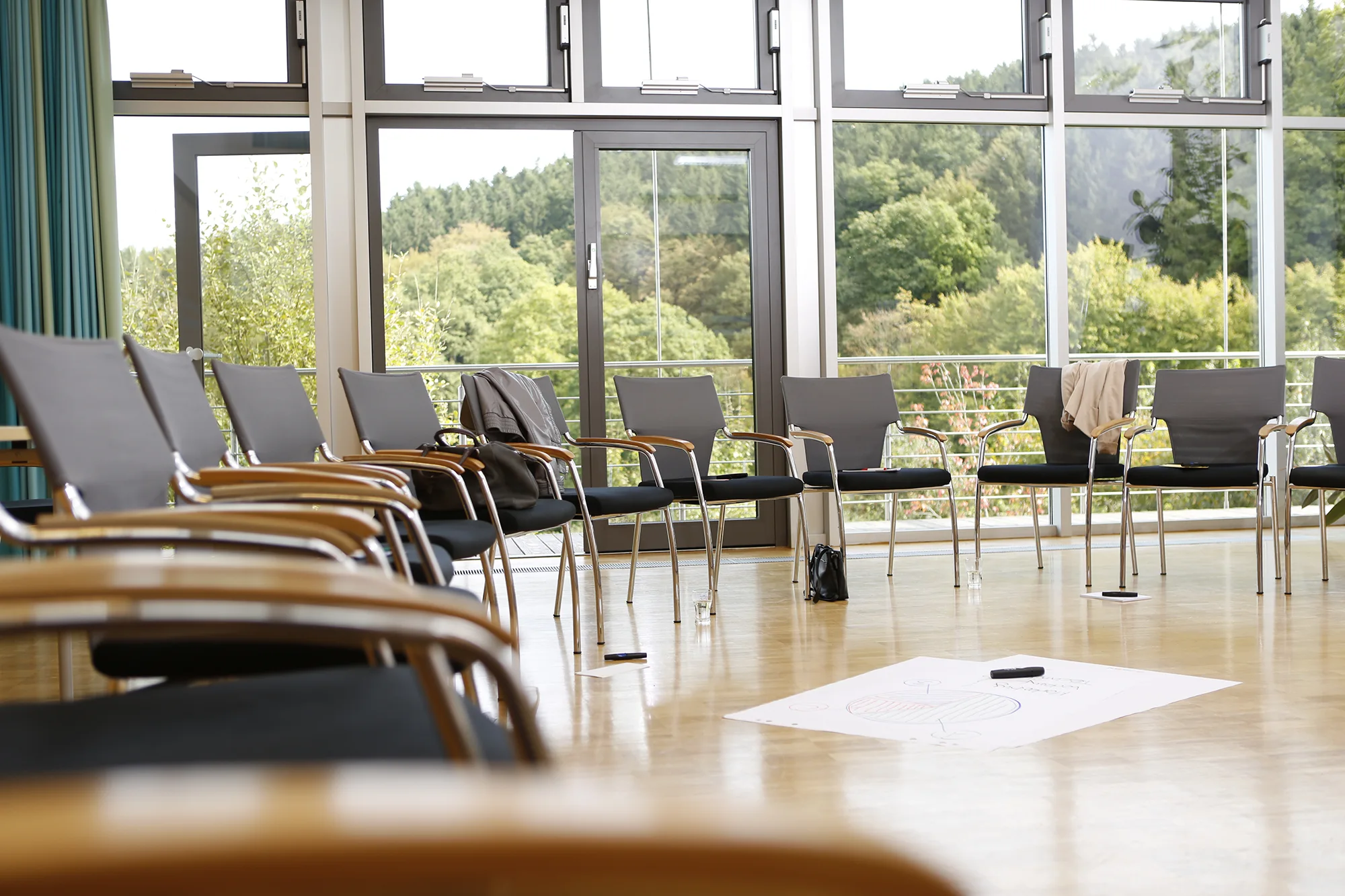Raum mit Blick ins Grüne durch eine komplette Fensterfront, auf Laminatboden steht ein Kreis aus Stühlen, in der Mitte ein Flipchart-Blatt mit Stift auf dem Boden