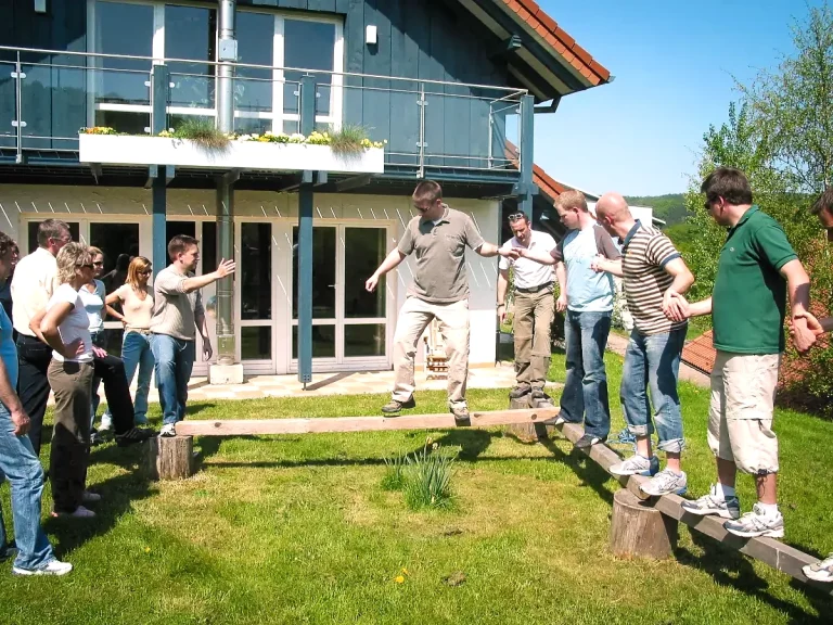 Eine Gruppe Menschen balanciert auf Holzbalken, die etwa 20 Zentimeter über dem Rasen aufgebockt sind