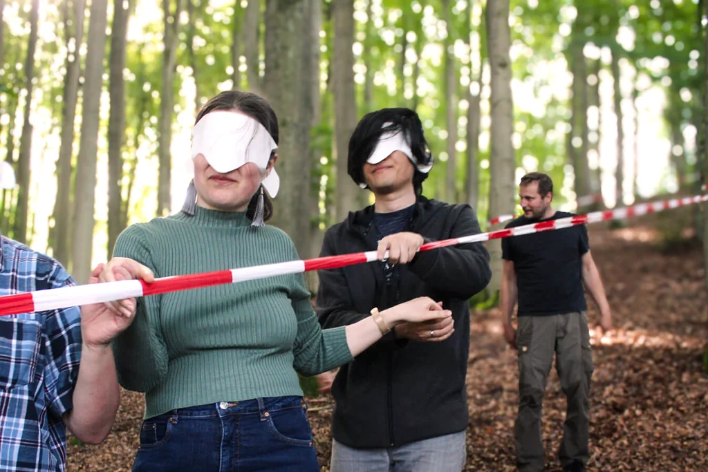 Eine Gruppe Menschen wir mit verbundenen Augen im Wald einen kleinen Abhang hinabgeführt, zur Orientierung dient ein Absperrband