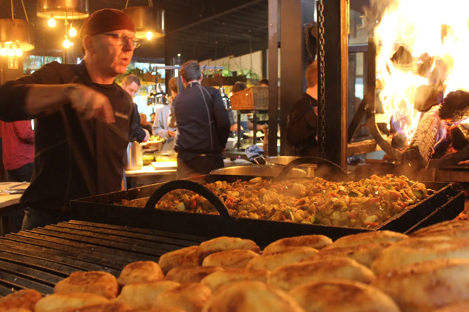 Ein Koch wendet Gemüse, das in einer großen flachen Eisenpfanne auf dem Grill gegart wird