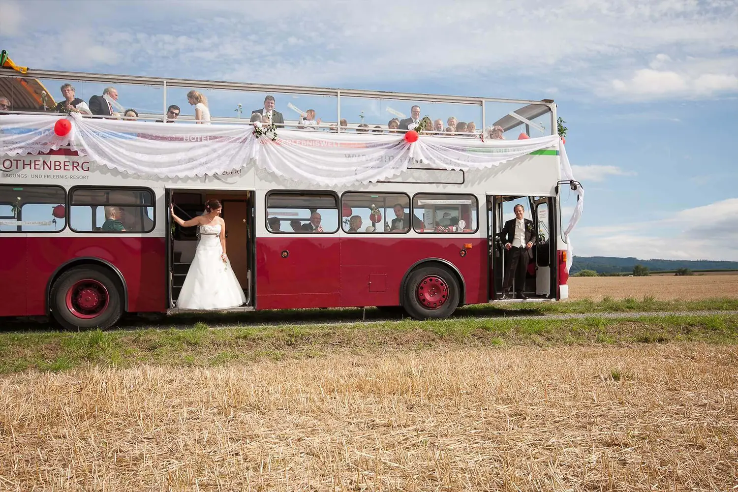 Ein Hochzeitspaar steht in den Einstiegstüren eines Oldtimer-Doppeldecker-Busses, der die Hochzeitsgesellschaft transportiert.