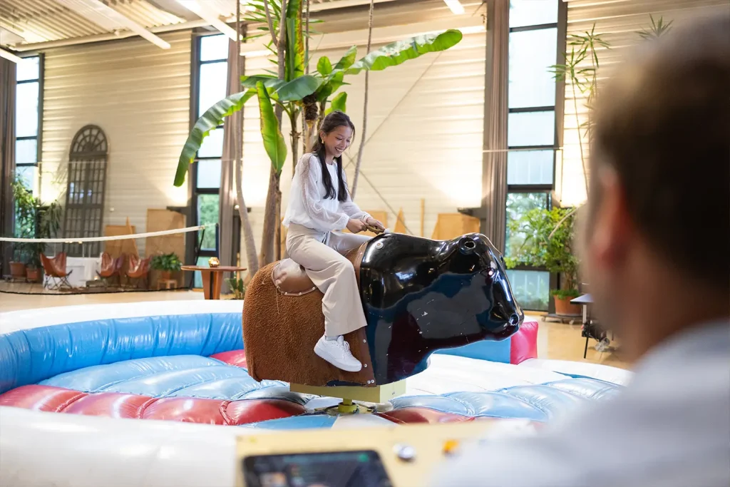 Eine junge Frau sitzt lachend auf einem Metallbullen beim Bullenreiten in einer Halle