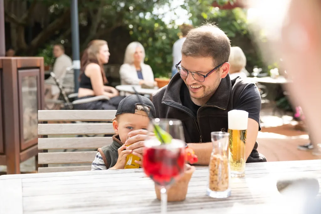 Ein Mann sitzt mit seinem kleinen Sohn in einem Biergarten und ist ihm beim Trinken aus einem Glas behilflich