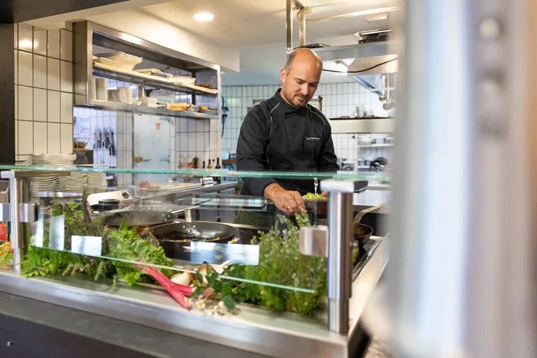 Ein Koch bereitet eine Speise in einer Pfanne zu, man kann ihm dabei über eine Glastheke hinweg zusehen