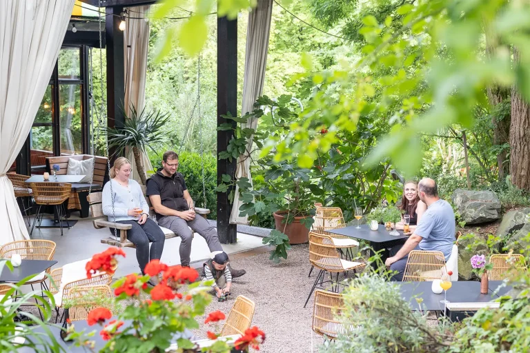 Eine Gruppe Menschen auf einer Terrasse, die von vielen grünen Pflanzen umgeben ist. Ein Paar sitzt gemeinsam auf einer großen Holzschaukel.