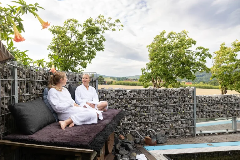 zwei Frauen in Bademänteln sitzen im Aussenbereich auf einer erhöhten Bank und unterhalten sich