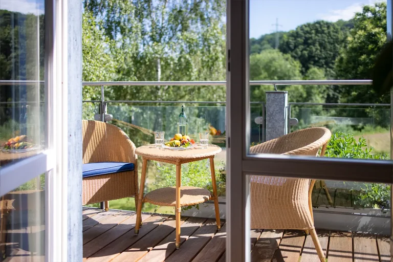 Ein Balkon mit Tisch und zwei Stühlen aus Korbgeflecht. Die Balkonwand ist aus Glas. Im Hintergrund grüne Bäume und blauer Himmel.