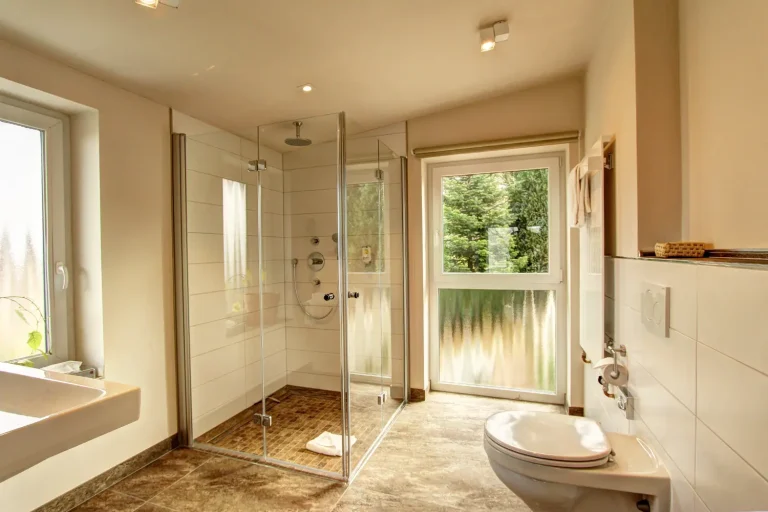 Badezimmer in hellen Farben mit großer Dusche, Toilette und Waschbecken, sowie einem großen Fenster