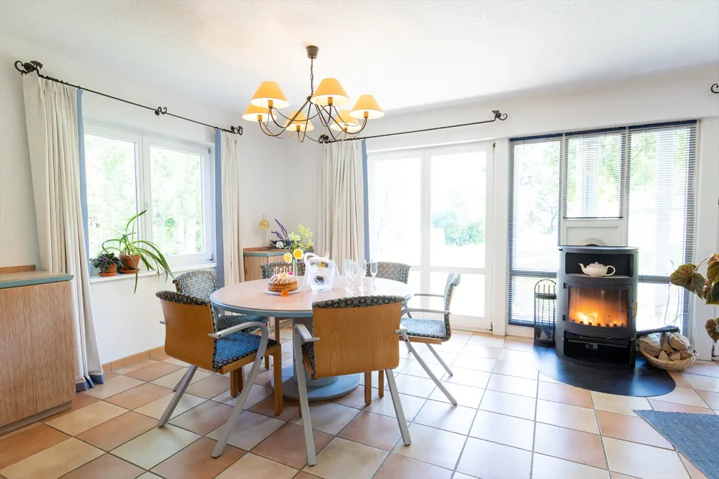 Runder Esstisch mit sechs Stühlen in einem Wohnraum mit brennendem Holzofen und großen Fenstern