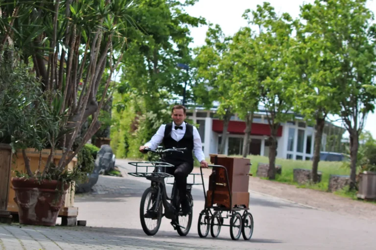Ein Mann in festlicher KLeidung mit Fliege, der auf einem Fahrrad fährt und dabei eine Drehorgel auf Rädern zieht