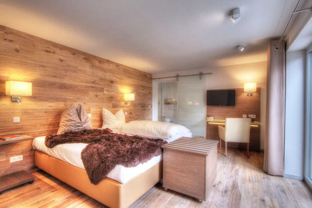 Hotelzimmer in Holztönen mit großem Doppelbett, Schreibtisch, Fernseher und Glastür, die zu einem Badezimmer führt