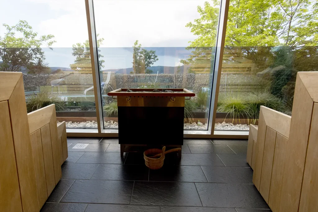 Eine große Fensterfront, durch die man ins Grüne schaut. Davor steht ein Saunaofen, rechts und links reihen sich Saunabänke aus Holz auf