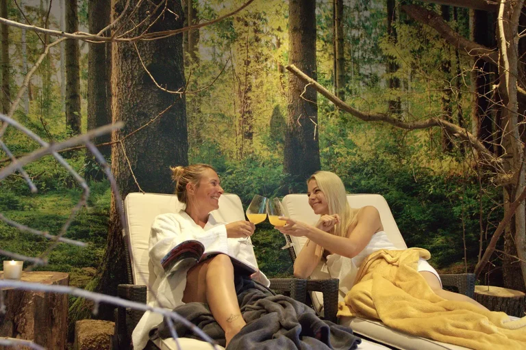 Zwei Frauen in Bademänteln und Handtüchern gekleidet liegen auf einer Liege und prosten sich mit einem Getränk zu