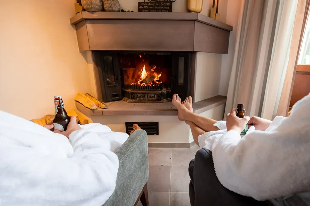 Ein Kamin mit offenem Feuer, davor sitzen zwei Menschen in Bademänteln mit Getränken in der Hand und hochgelegten Füßen