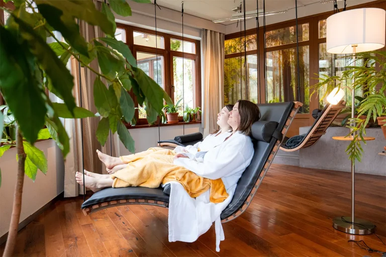 Zwei Frauen in Bademänteln liegen auf einer Liege, die ergonomisch geformt ist und von der Raumdecke abhängt