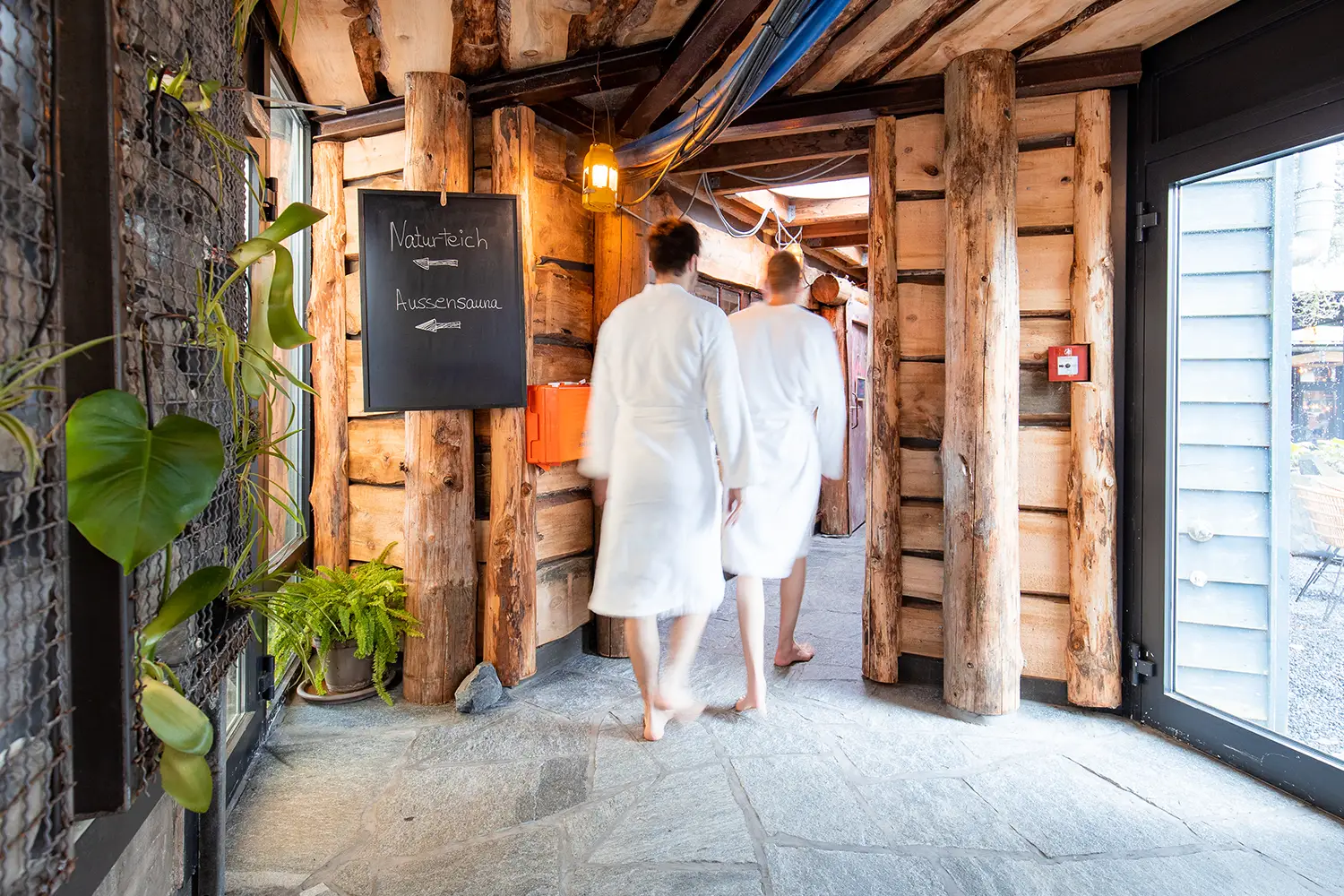 Zwei Männer in Bademänteln, die einen Gang entlanggehen, der wie in einem Bergwerkstollen an den Wänden und der Decke mit Holzbalken verkleidet ist