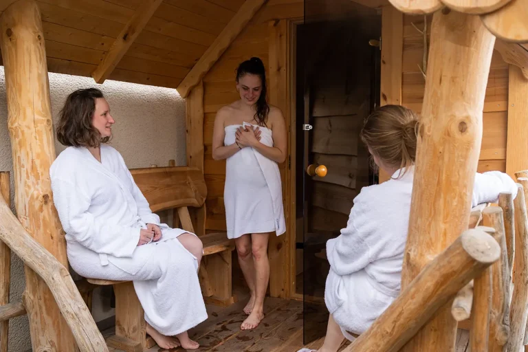 drei Frauen in Bademantel und Handtuch gekleidet stehen vor einer Blockhaussauna und unterhalten sich