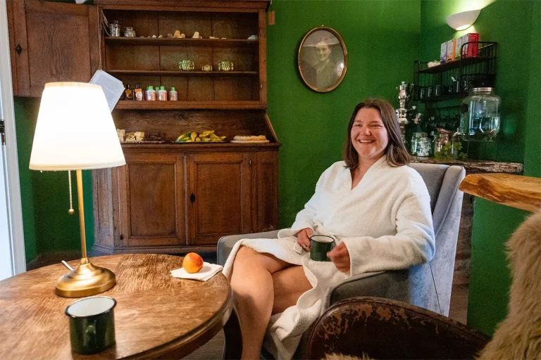 Eine Frau mit Bademantel bekleidet sitz mit einer Tasse in der Hand auf einem Sessel vor einem kleinen Tisch