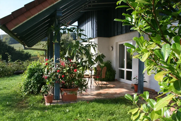 geflieste Terrasse unter einem Schrägdach mit vielen blühenden Kübelpflanzen und Blick ins Grüne