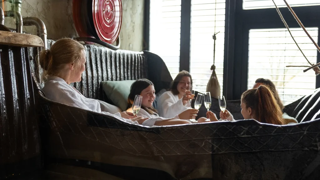 Vier Frauen in Bademänteln sitzen gemeinsam in einer großen Wanne und halten Sektgläser in der Hand