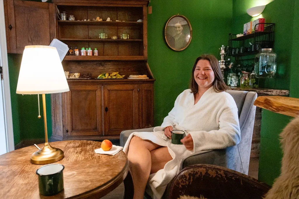 Eine Frau im Bademantel sitzt in einem Sessel mit einer Tasse in der Hand und lacht