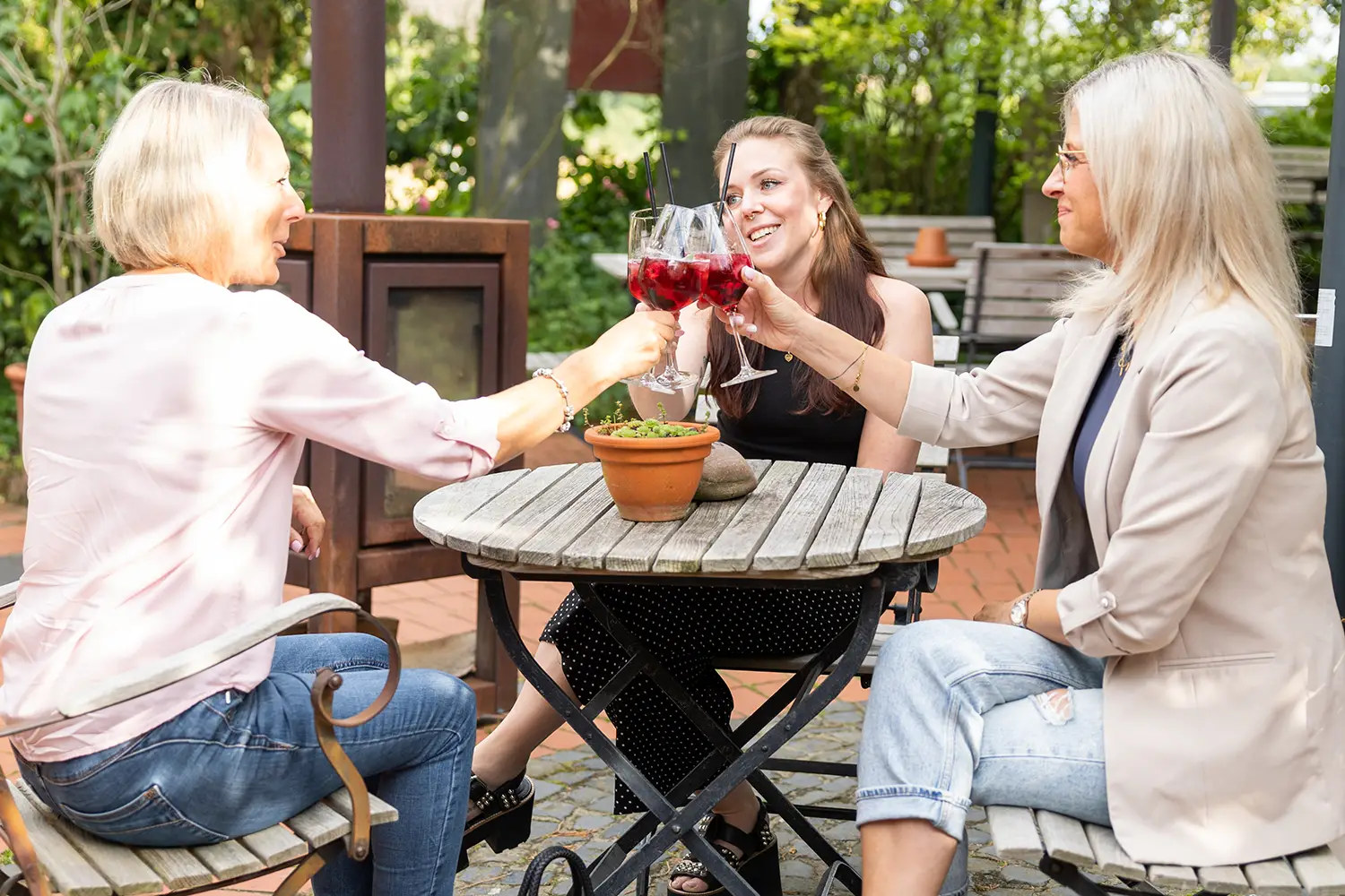 Drei Frauen sitzen in einem Biergarten und prosten sich mit Cocktails zu