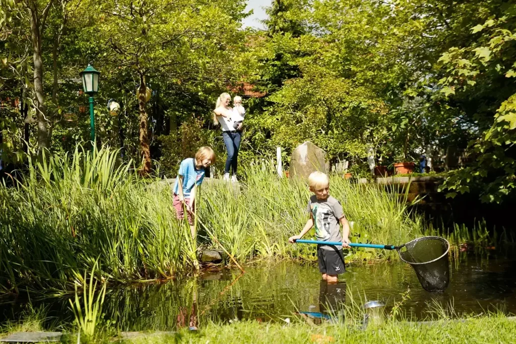 Eine Frau mit drei Kindern an einem Teich, die Kinder fangen etwas mit einem Käscher
