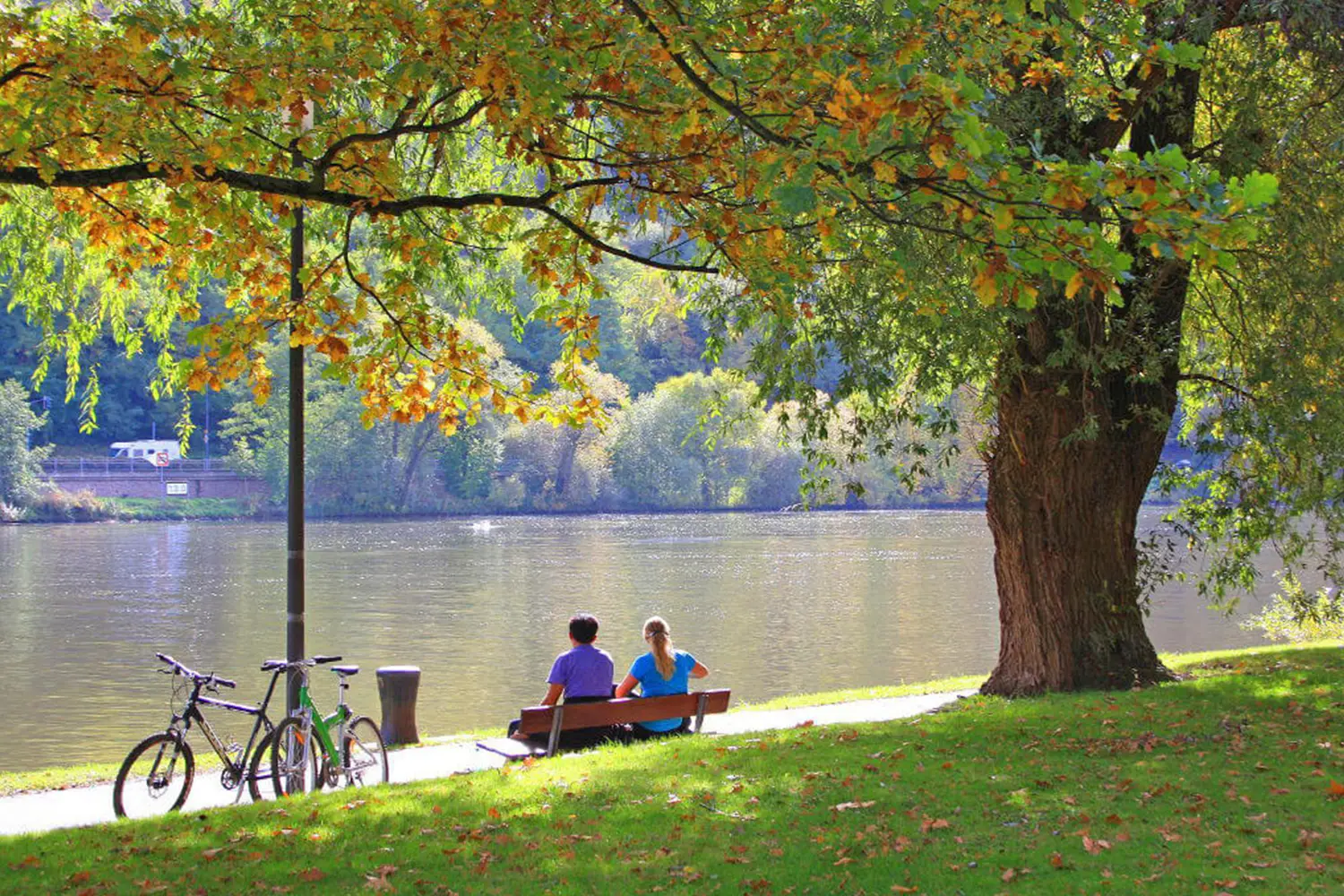 Zwei Menschen sitzen auf einer Bank an einem Fluss, daneben zwei Fahrräder